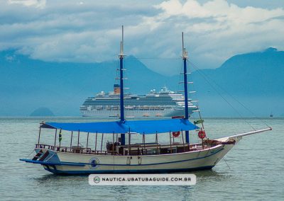 Em plano aberto e com a Serra do Mar em terceiro plano e a escuna no primeiro, destaca-se o grande navio turístico entre elas