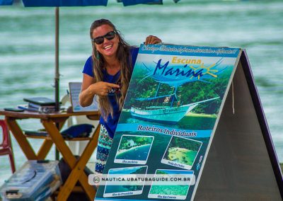 Apontando para a placa com sorriso estampado, Jennifer é exatamente assim, sempre sorridente ao falar de passeios de escuna em Ubatuba
