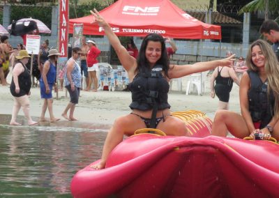Duas jovens mulheres sorrindo a bordo de banana boat duplo com a tenda da FNS Turismo ao fundo na Praia do Lázaro