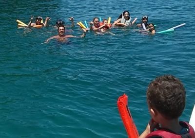 Pessoas praticando flutuação com espaguetes observadas por criança prestes a pular na água com colete salva vidas durante passeio de lancha em Ubatuba